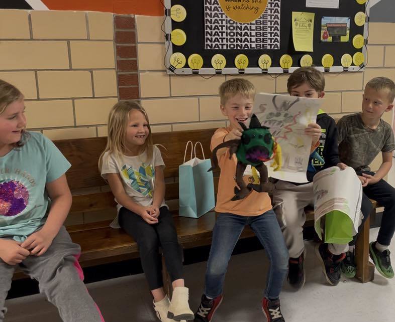 Five children sit on a bench; one holds a book and a green puppet. Others hold books, bags, and a puppet. A bulletin board is on the wall.