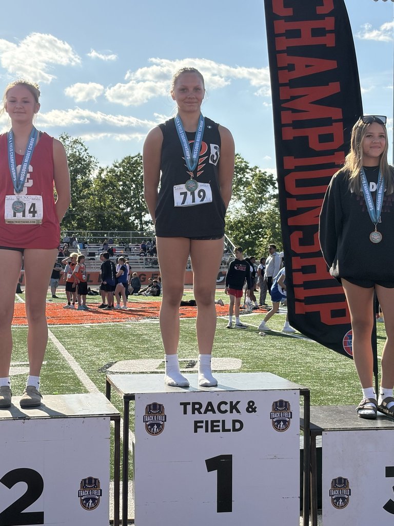 Three individuals stand on a podium with signs for 1st, 2nd, and 3rd place. All wear medals and athletic wear. Behind them is a grassy field with a blurred banner.