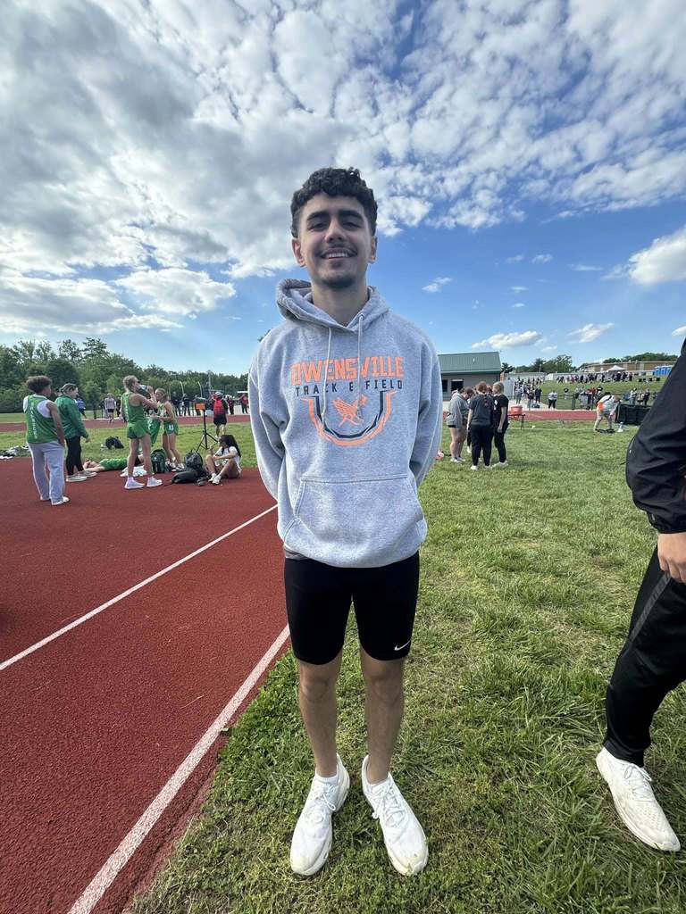 Man standing on grass near a track with a red running lane, wearing a gray hoodie, black shorts, and white shoes.