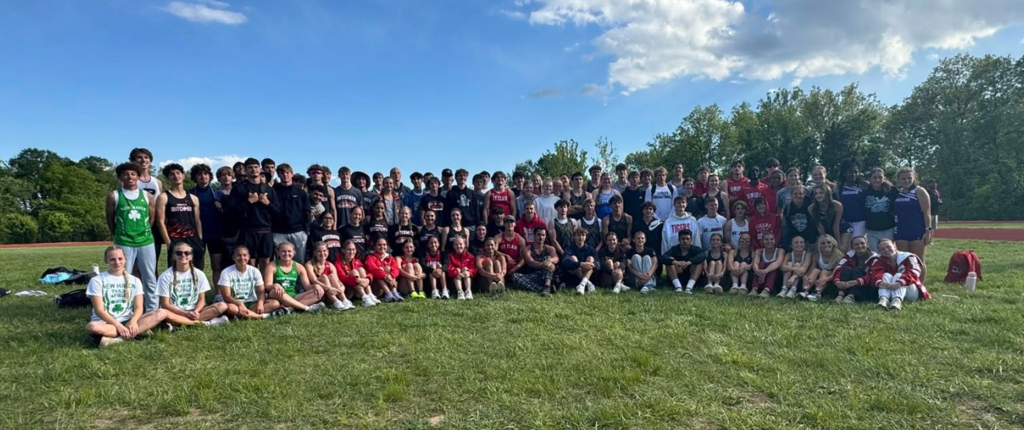A large group of people in sports attire pose on a grassy field with trees in the background.