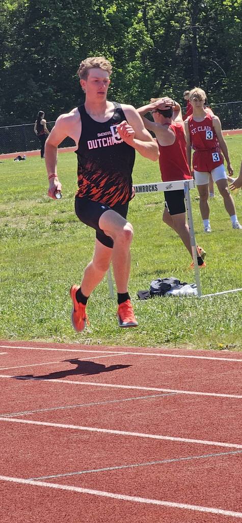 Man jumping hurdles with number 56 on shirt. Background shows grassy field with trees and people.