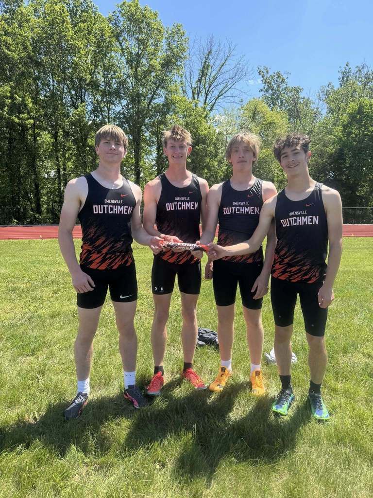 Four athletes in matching jerseys hold a silver trophy on a grassy field. Trees are in the background.