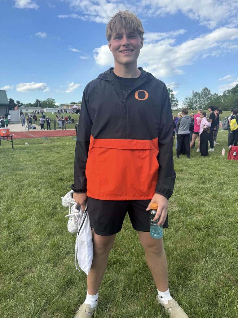 A young man wearing black and orange athletic wear stands on a grassy field, holding a water bottle.