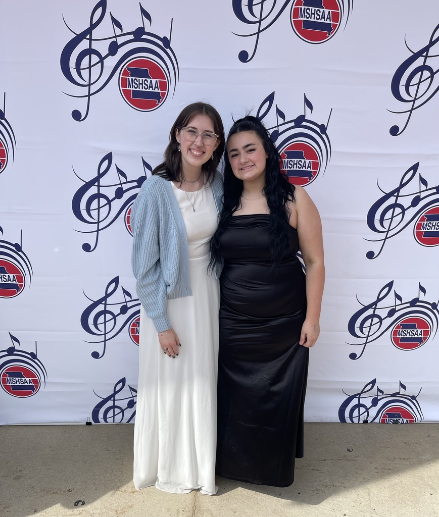 Two women, one in a white dress and the other in black, standing next to each other in front of a backdrop with musical notes.
