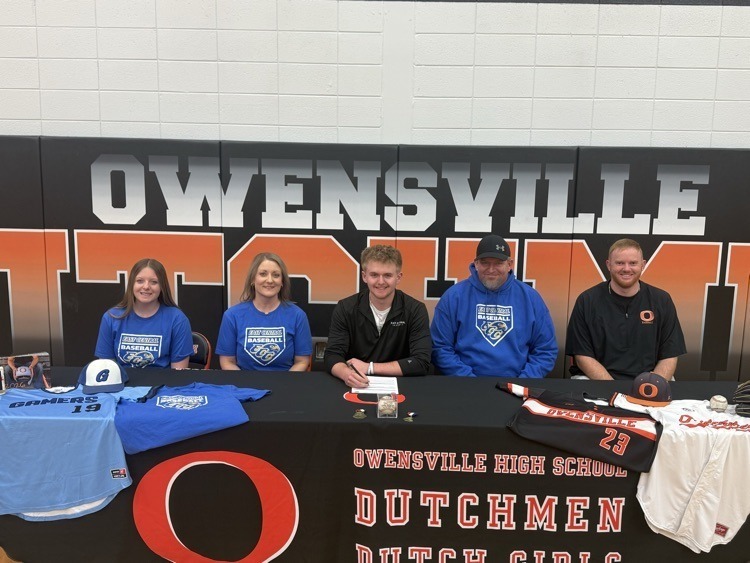 Five people in blue and black shirts pose at a signing table. The table displays blue and white shirts and a large O letter.