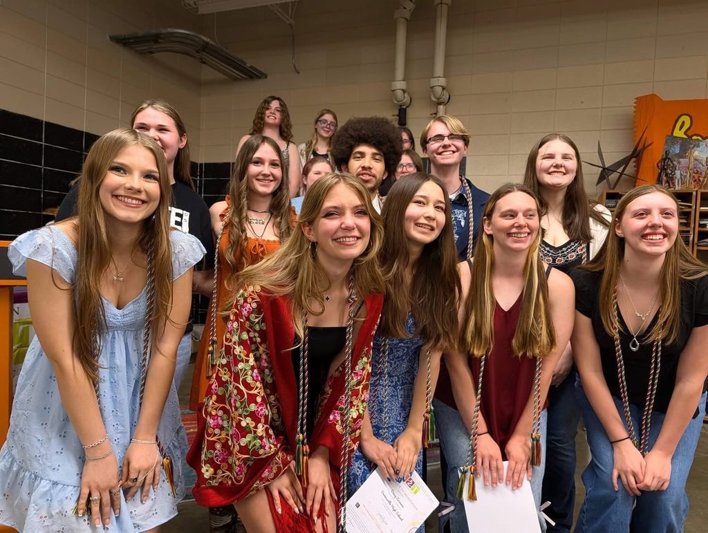 A group of young people stand together, smiling. Most wear casual clothes. One holds a piece of paper.