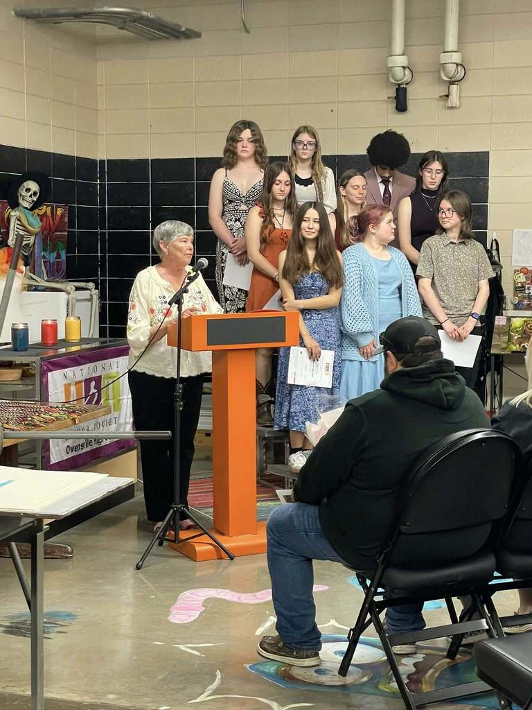 Several people stand at a podium; a woman speaks into a microphone. Behind them, others sit or stand in a room with a tiled wall.