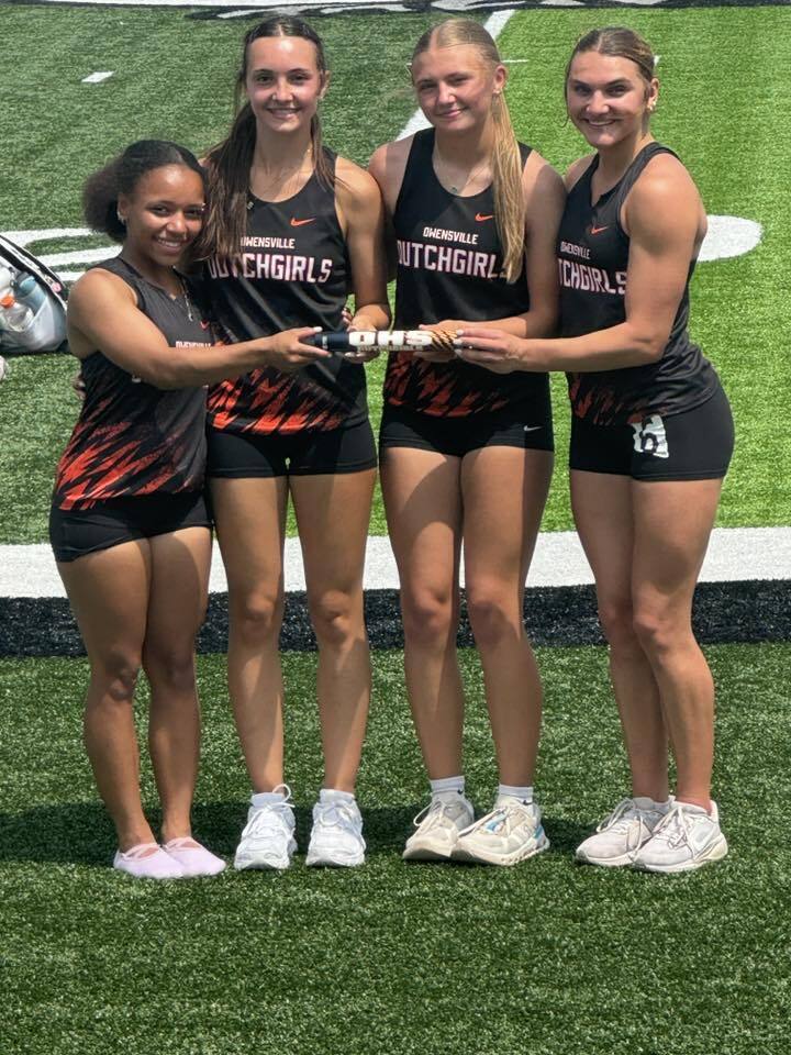 Four young women stand on a field in matching athletic uniforms, holding a trophy. They are smiling and appear to be posing for a photo.