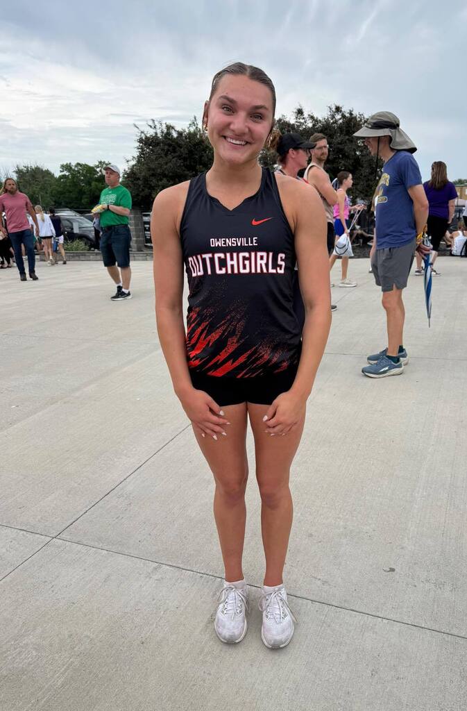 A young woman in a sports uniform stands on concrete with several people in the background.