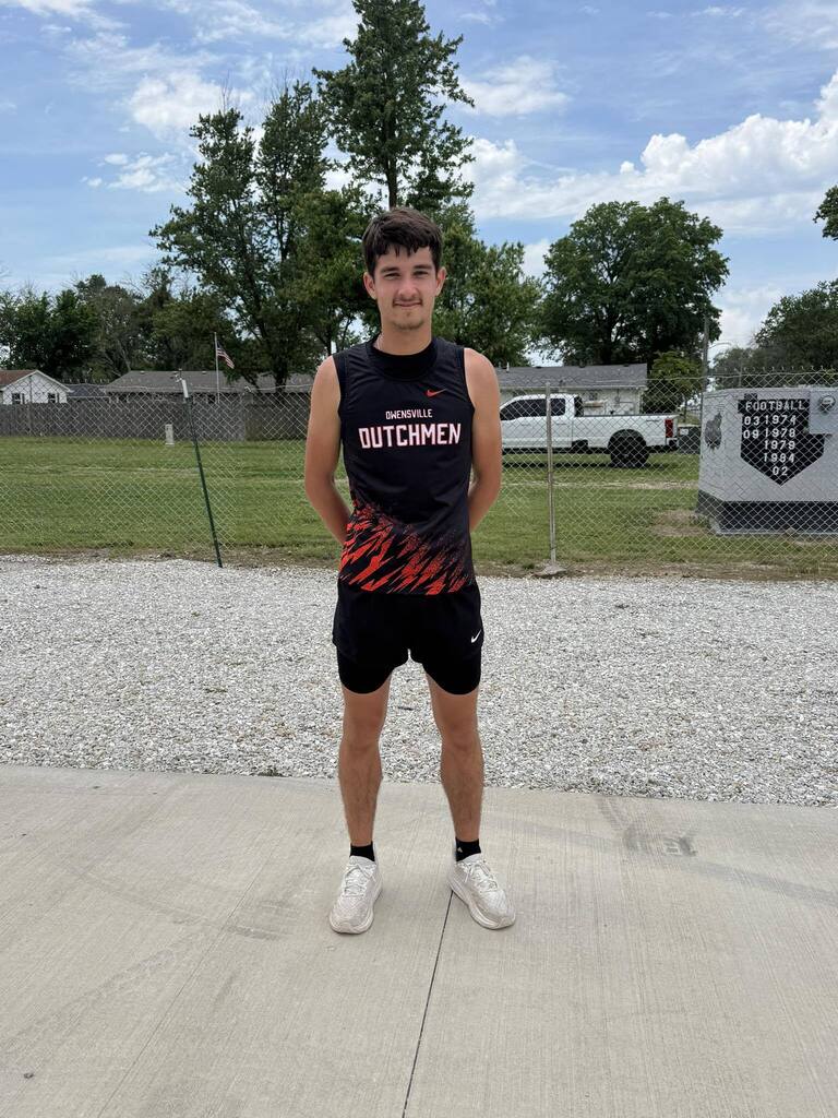 A man in athletic wear stands outside. Behind him is a grassy area with a fence and parked vehicles.