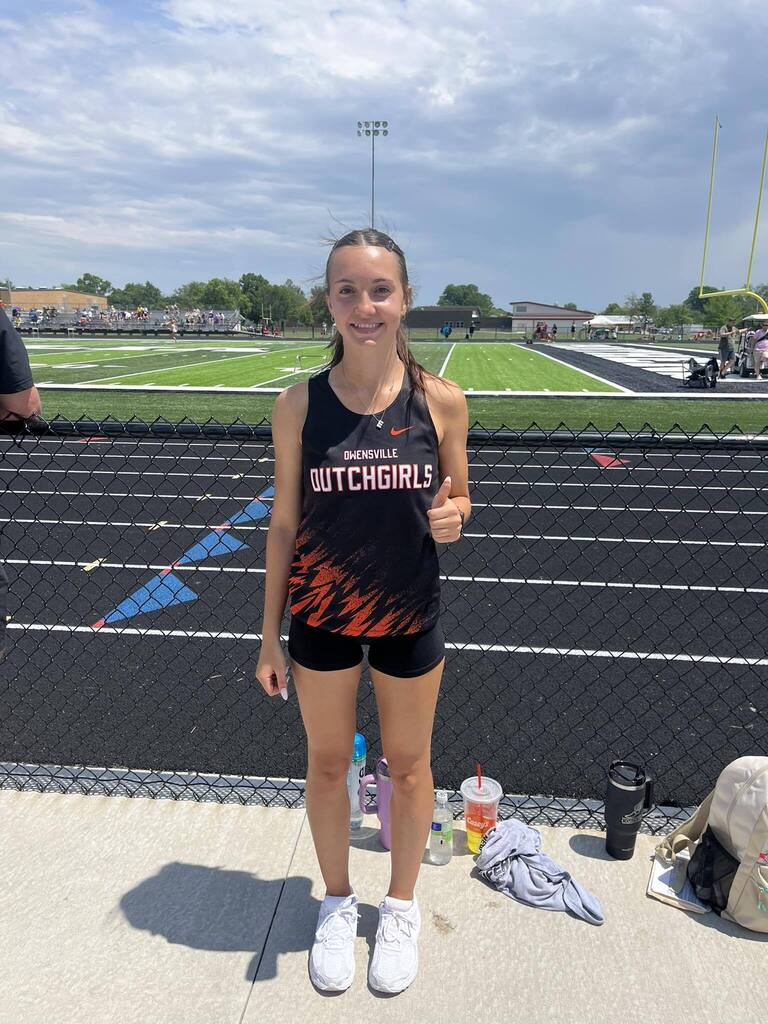 A girl in a Dutchgirls jersey and shorts poses with a thumbs-up on a track field.