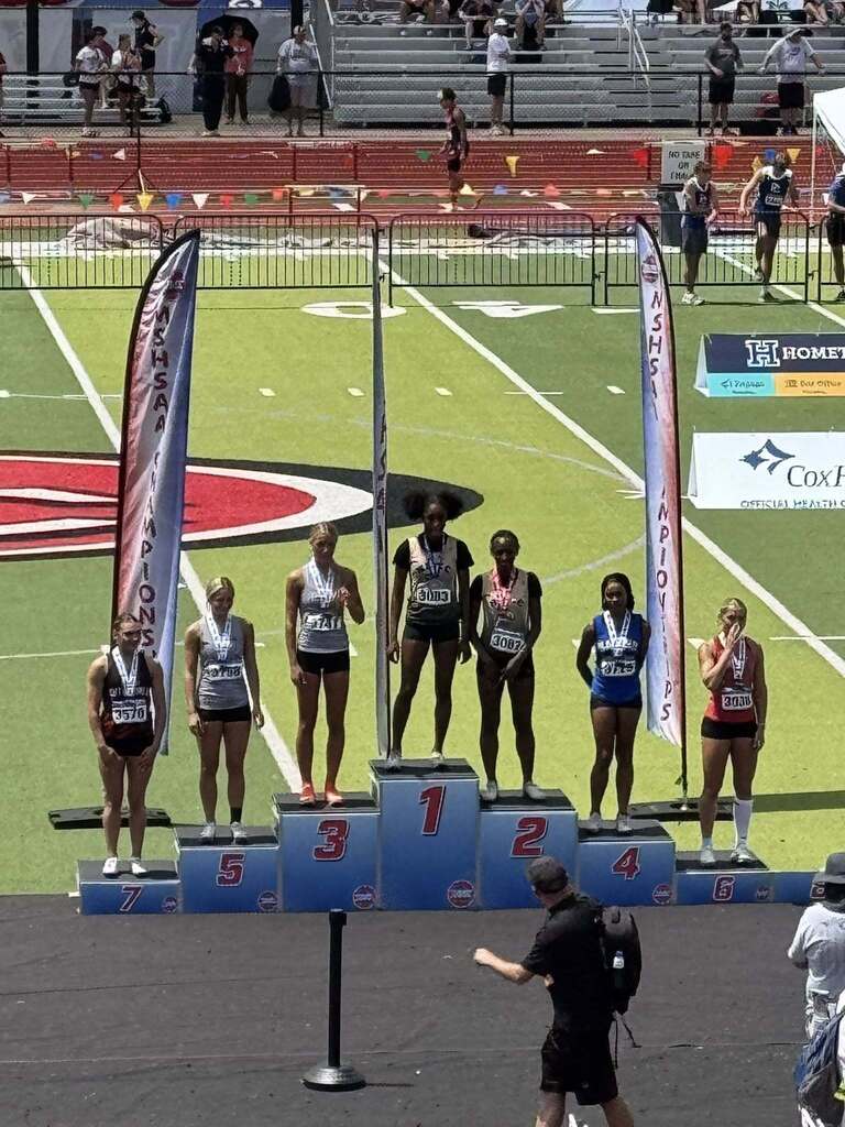 Athletes on a track field stand on a podium with numbers 1 to 5. Two banners are in the background.