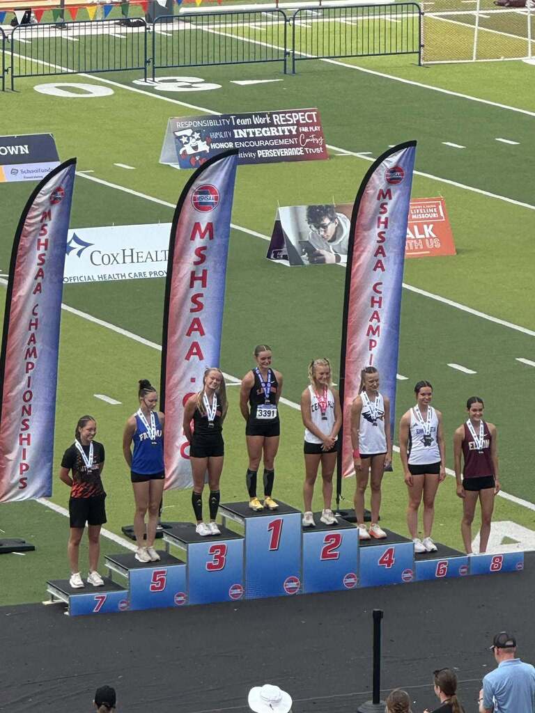 Athletes stand on a podium with numbers 1 to 5. Banners are behind them with sponsors and text. They wear athletic uniforms.