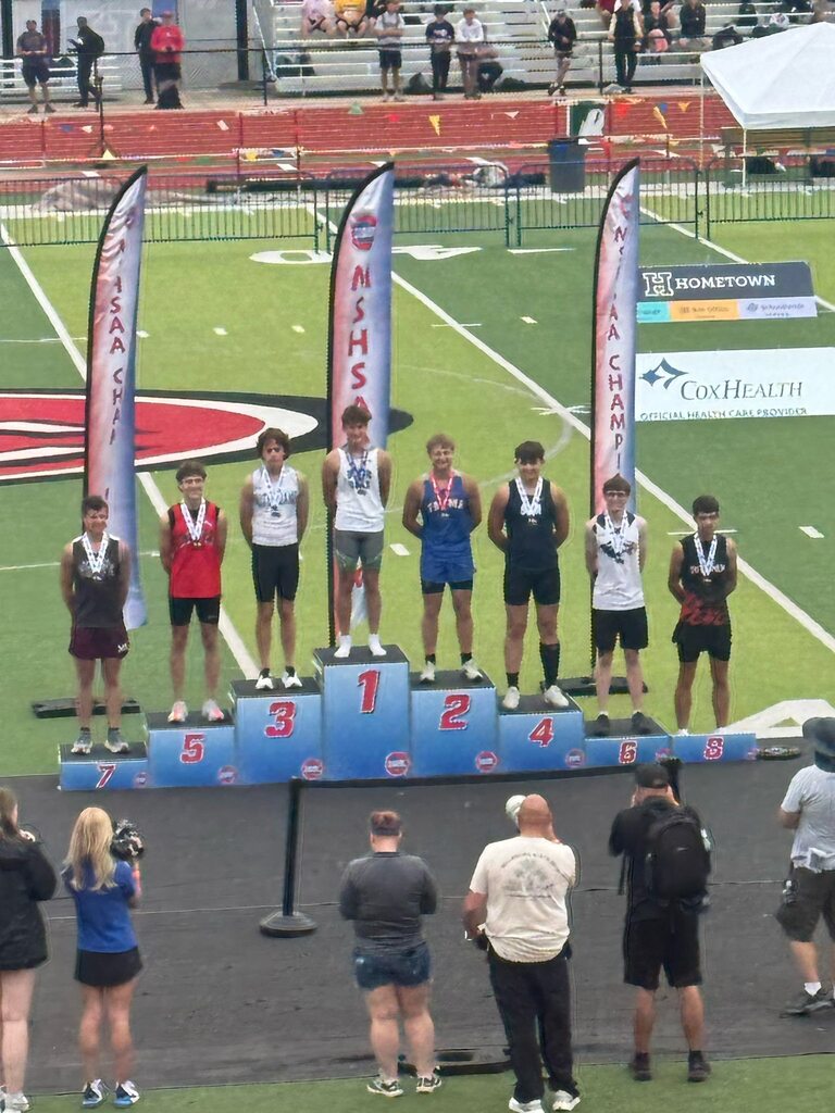 Seven people stand on a podium with numbers. A crowd watches from the sidelines of an athletic field.