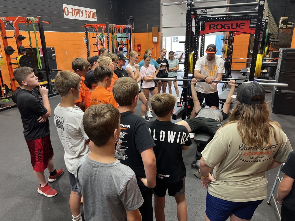 Group of young people observing a man lift weights in a gym. Bright lights and gym equipment visible.