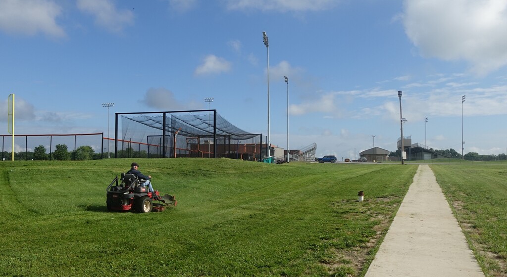 A person operates a lawn mower on a grassy field with a batting cage, a path, and poles in the background.
