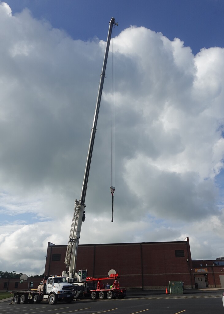 A tall crane extends upward, attached to a white truck. A red building is in the background, and the sky is cloudy.