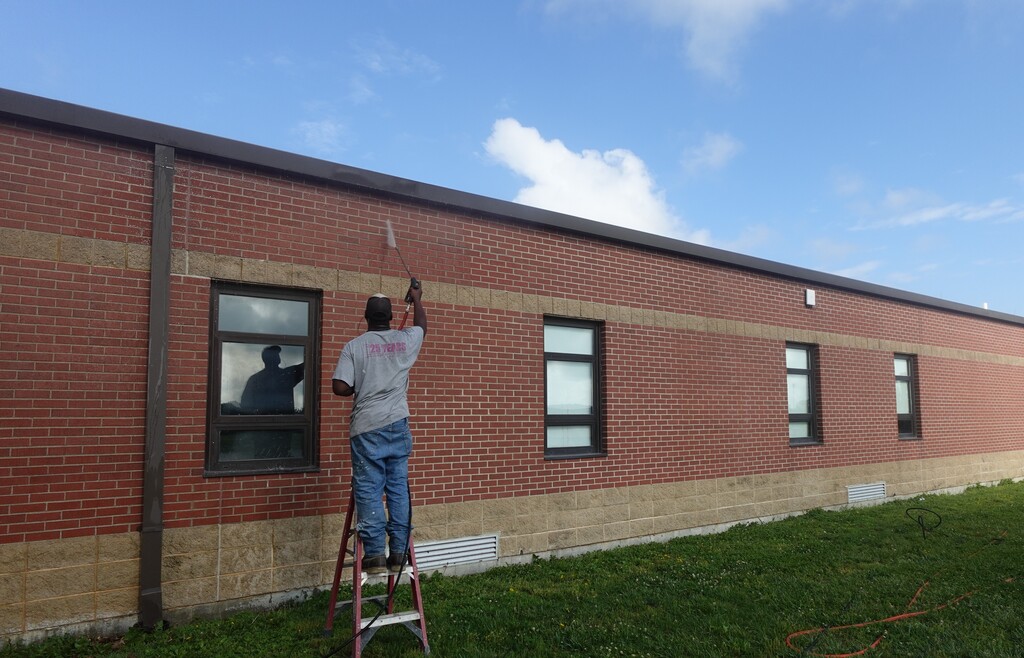 A person paints a brick building with windows. The building has a grassy lawn in front.