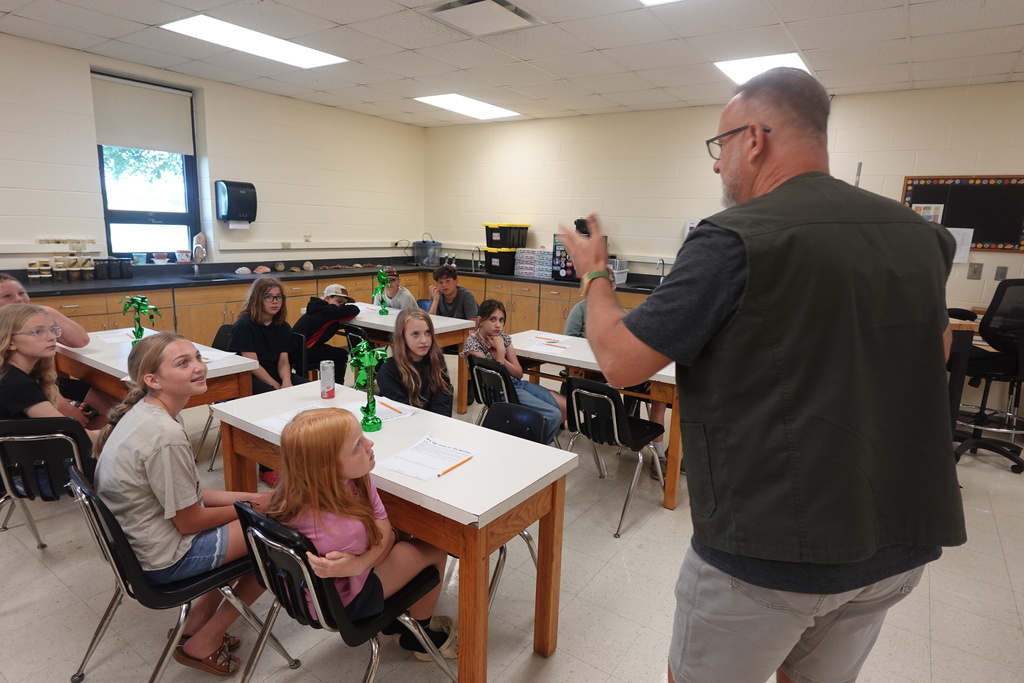 A classroom with students seated around desks. A man stands before them, holding a phone, possibly explaining something.