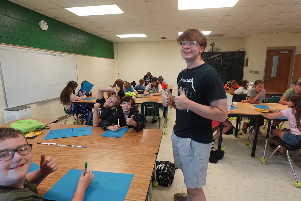 Students sit at desks in a classroom, one giving a thumbs up. A teacher stands next to them.