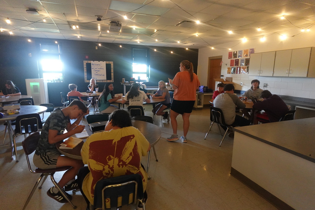 Group of students sitting at tables in a room. A person stands near the back. A wall has cabinets and pictures.
