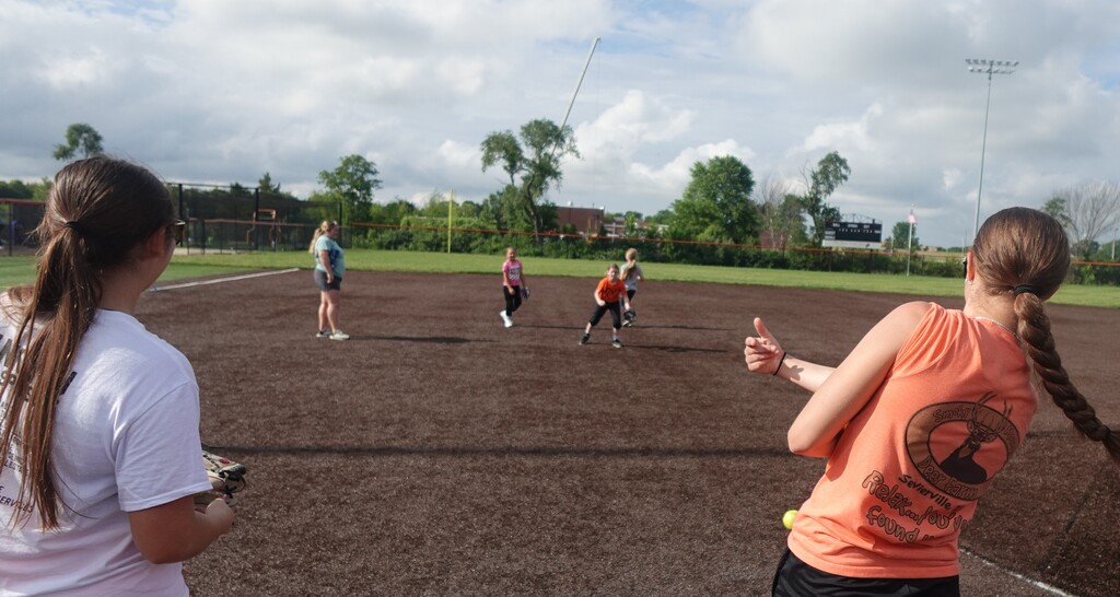 A group of people play softball on a field with a cloudy sky. A woman with a ponytail watches from the side.