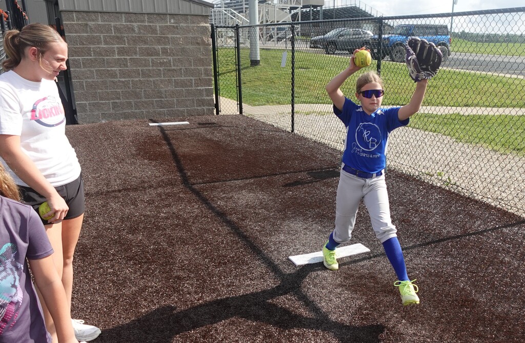 A girl in a blue jersey catches a ball at a baseball field, with two onlookers nearby.