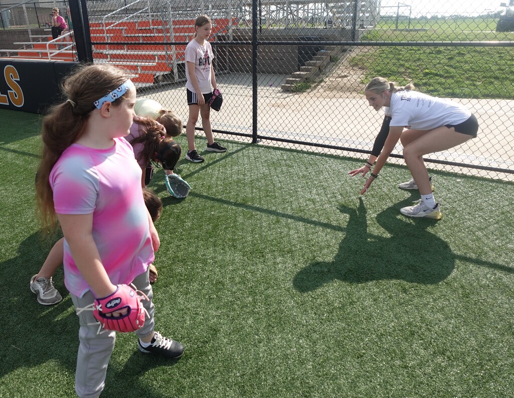 A group of young people play softball on a green artificial field. Some wear gloves and sneakers.