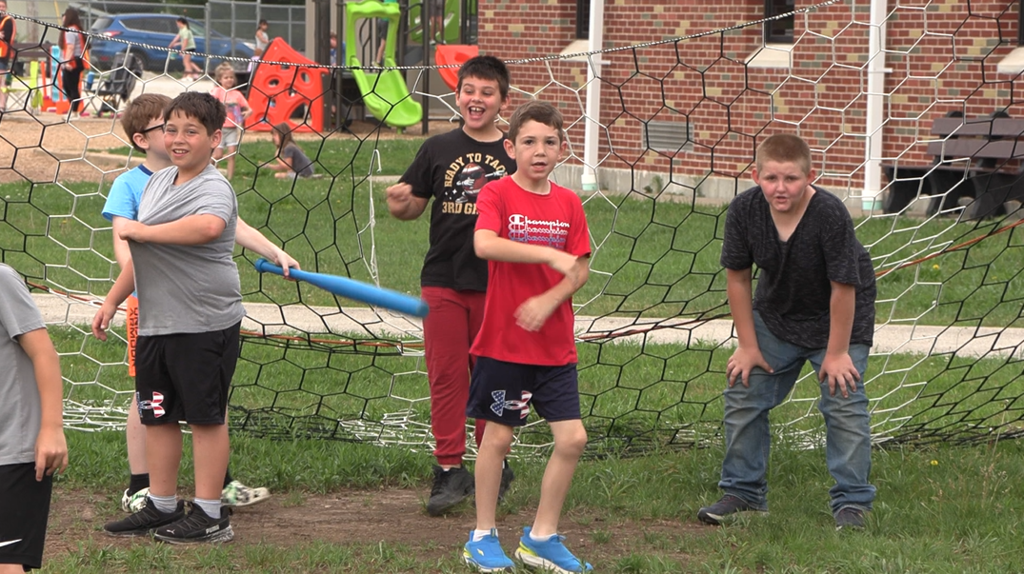 Six children standing outside with a soccer goal in the background. Three holding a bat, two in red pants.