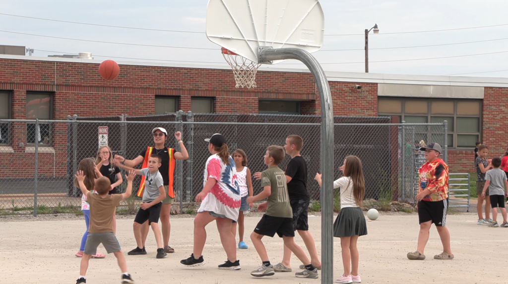 Several kids and adults on a basketball court, one person shoots basketball, some are walking.