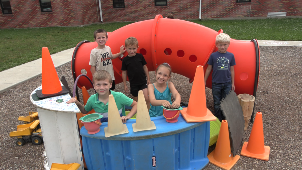Five children, smiling, pose in front of orange playground equipment and orange traffic cones in a park.