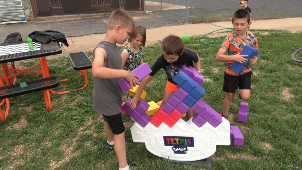 Four children on a grassy area with a large block structure, two boys holding blue blocks.