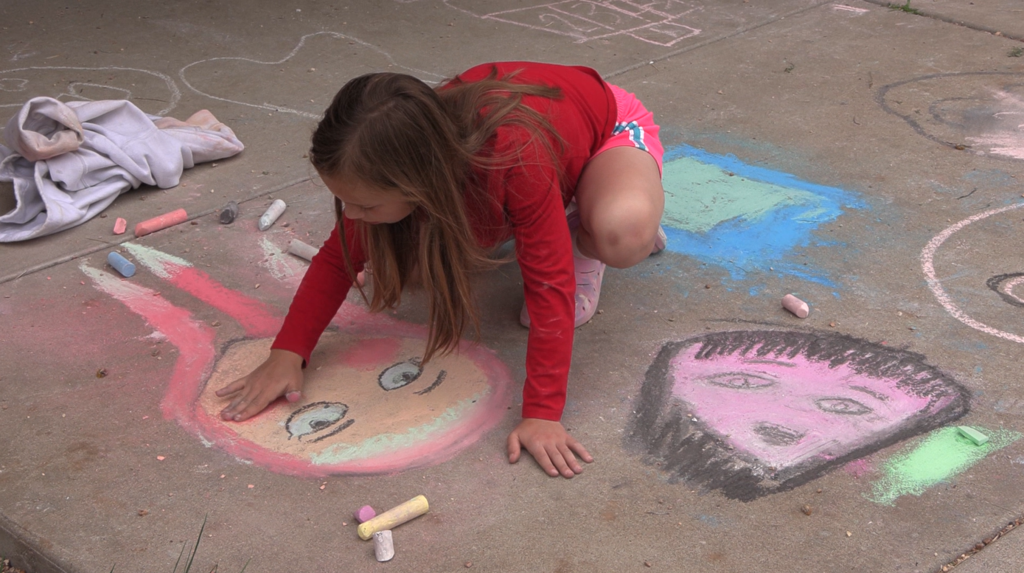 A girl kneeling on concrete, drawing a colorful face with chalk. Other chalk drawings are visible on the ground.