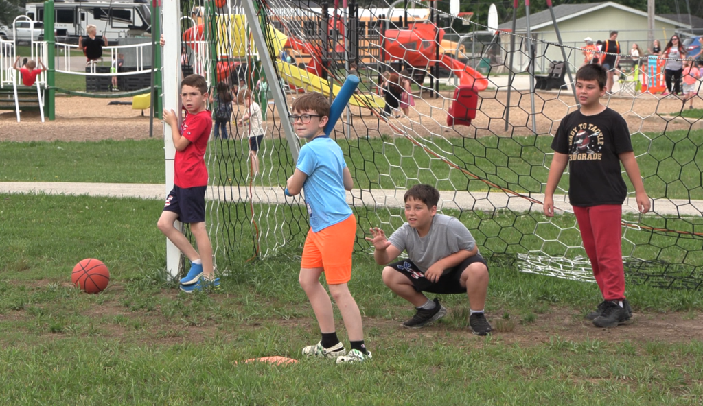 Four children play on a grassy field, one holding a bat and another a ball near a net.