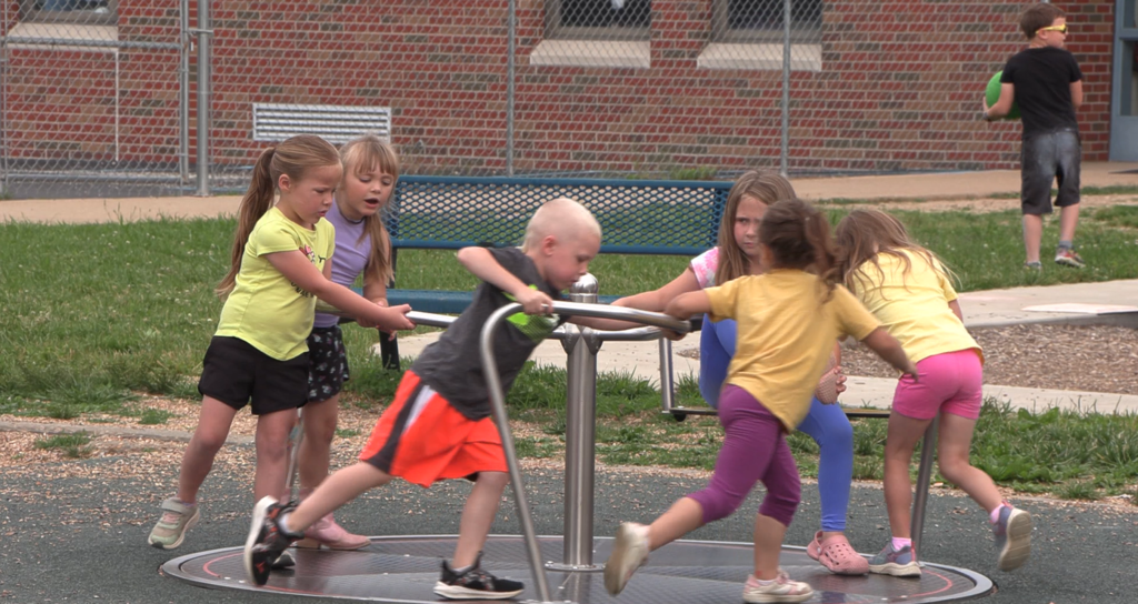 Five children play on a merry-go-round in a park; three in yellow and one in gray, orange, and pink.