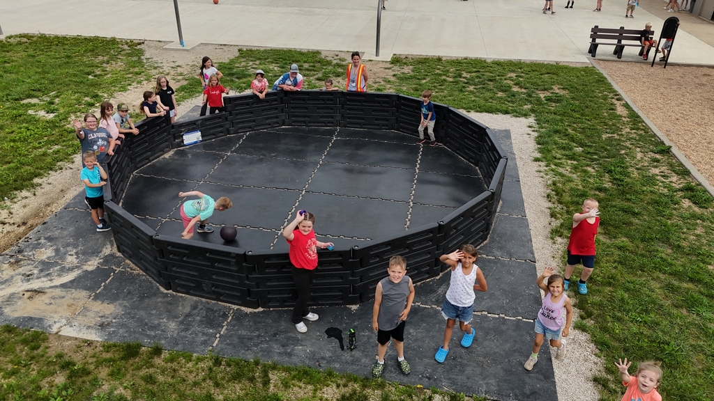 Children in various colored shirts and shoes play in a circular outdoor playground with black walls.