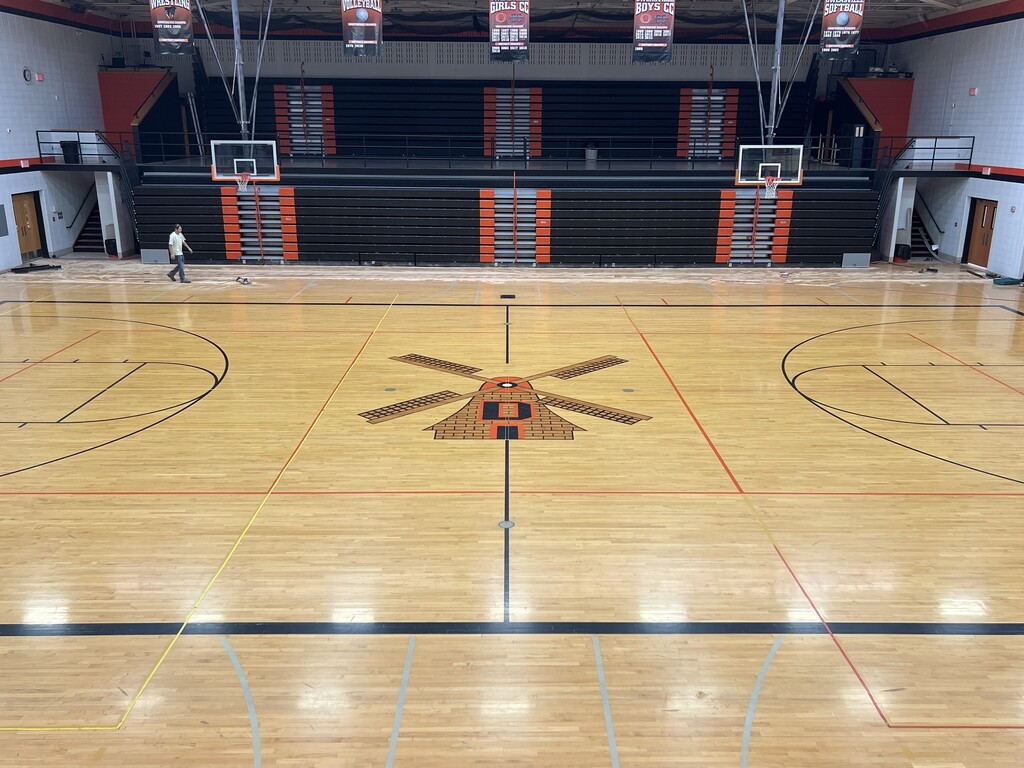 An indoor basketball court with wooden flooring, black borders, and a logo. Two people are walking, and the bleachers are empty.