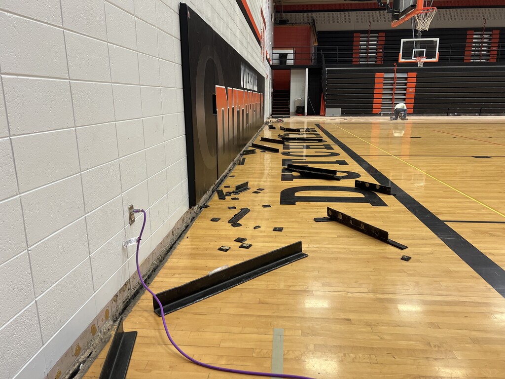 An indoor basketball court with a broken floor, a black "DRAG" sign, a basketball hoop, and wires on the wall.