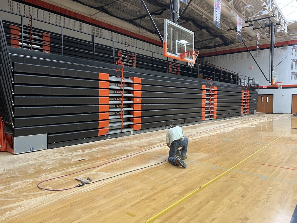 An individual kneels on a basketball court, cleaning the floor with a mop. The court is bordered by empty bleachers.