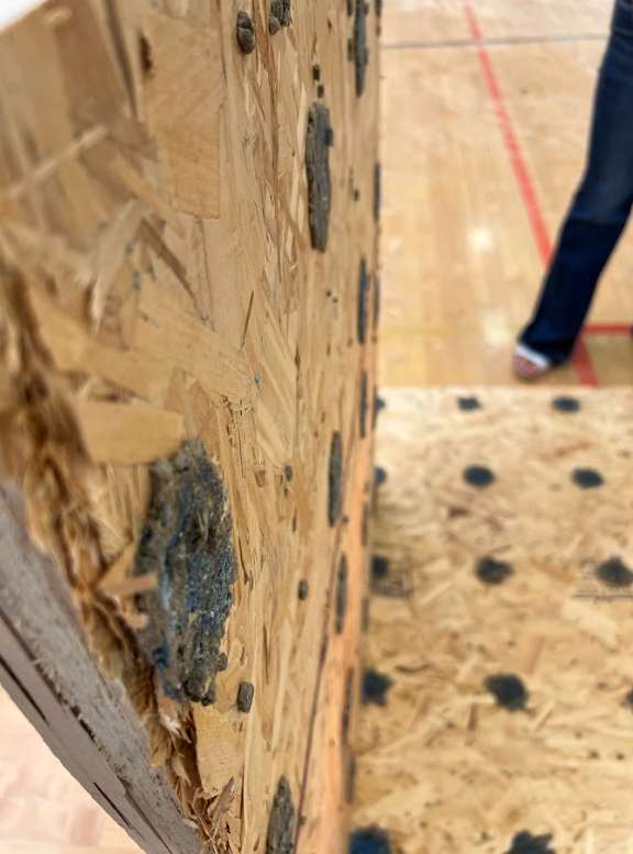 Close-up of a climbing wall in a gym. Brown wooden wall with textured surface, black climbing holds. Floor with red line.