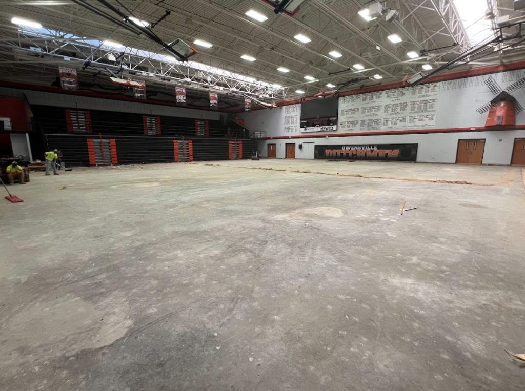 Large empty room with red and white walls, black benches, and ceiling lights. Workers are sweeping the floor.