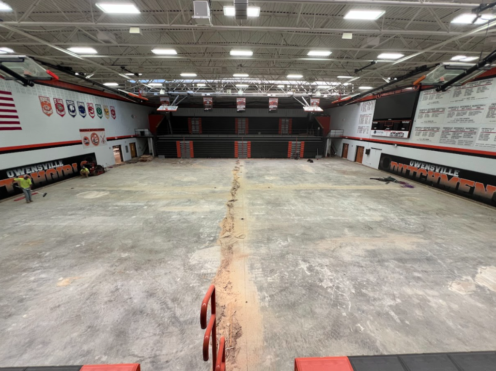 An empty sports hall with a high ceiling. The floor is concrete, and a crack runs through the middle.