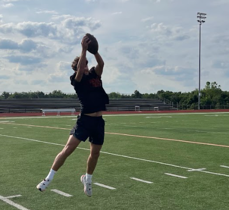 A person jumps mid-air to catch a football on an outdoor football field. Green grass, white lines, bleachers, and a tall pole are visible in the background.