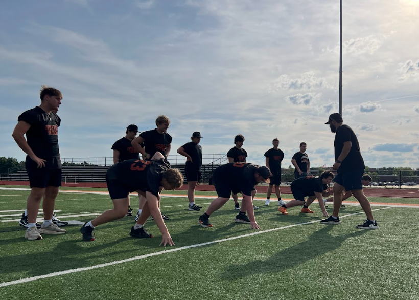 A group of athletes in uniform performs drills on a field, with one person bent over. A coach watches from behind.