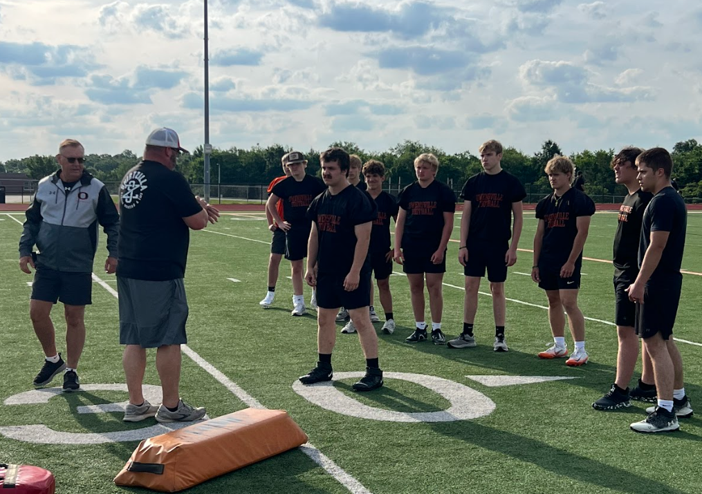 Nine athletes in black jerseys on a football field. One coach in a black shirt and cap talks.