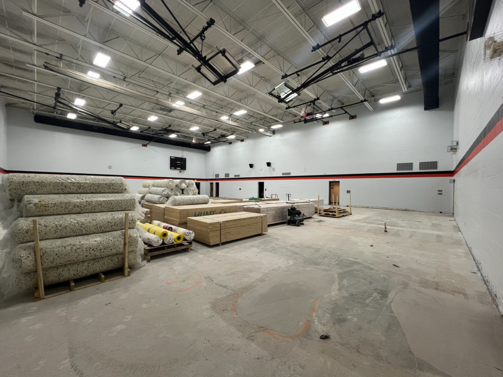 Interior of a large room with unfinished concrete floor and empty walls. Many materials are stacked on the floor.