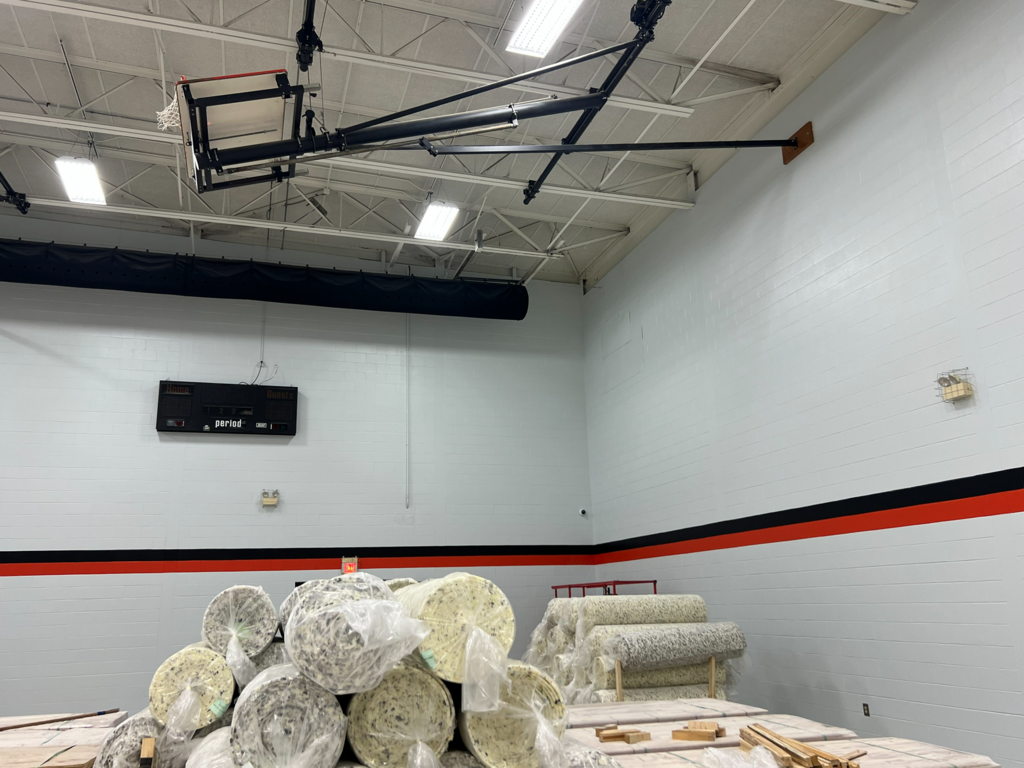 An indoor area with white walls, red and black stripes, a basketball hoop, and rolls of carpet.