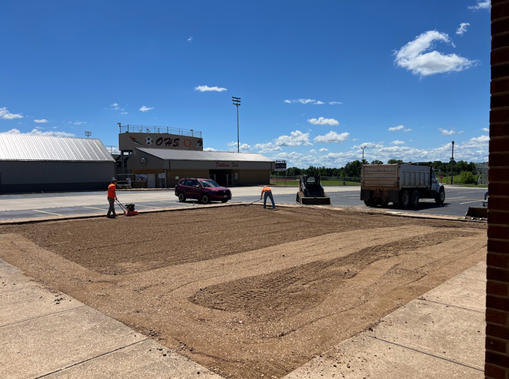 An outdoor area with a road and two workers. One person is using a machine. Three vehicles are parked.