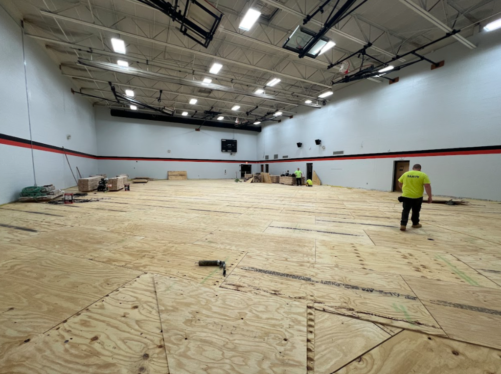 An indoor space with wooden flooring and exposed ceiling. A person in a yellow shirt is walking on the floor.