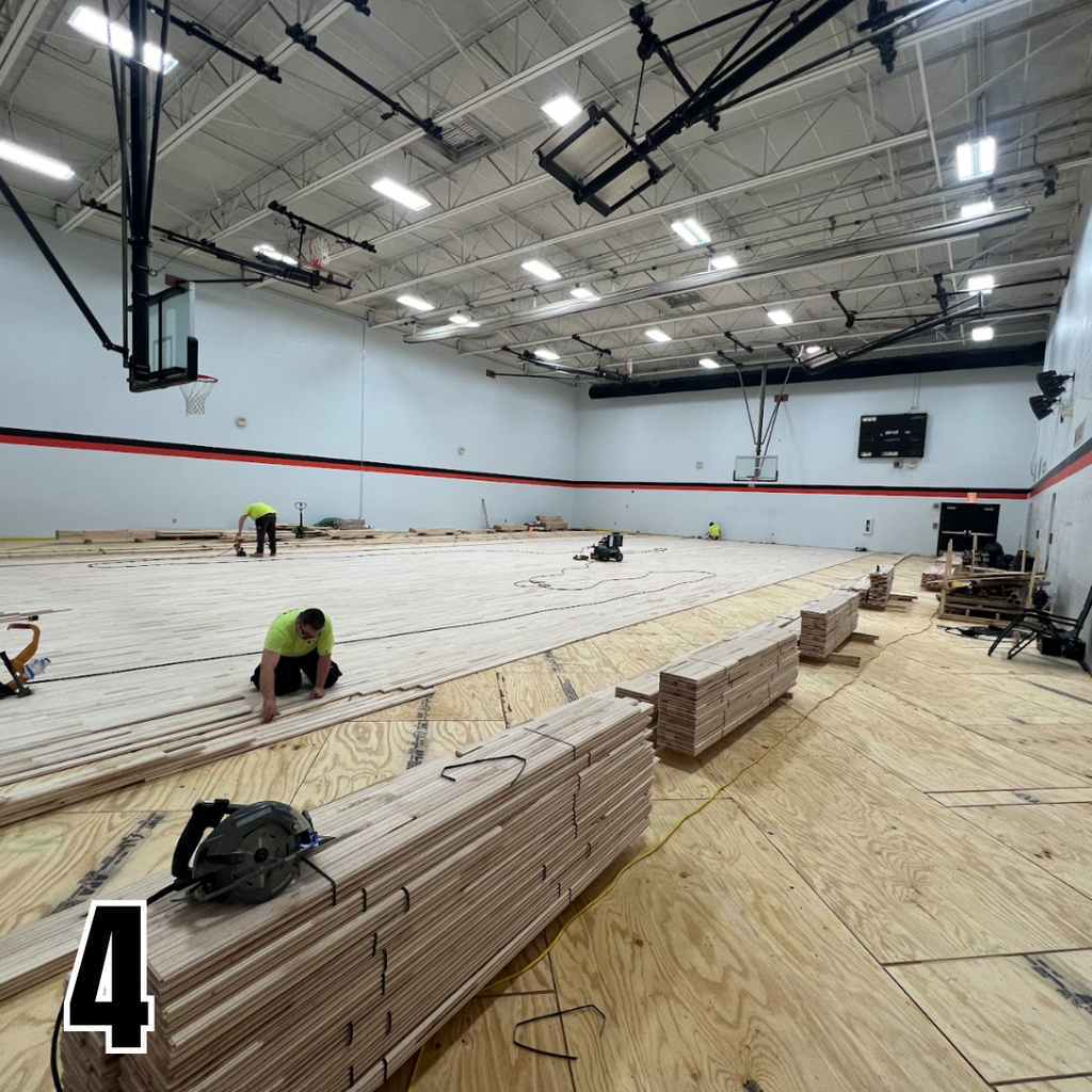 A basketball court under construction. A person in green works on the floor, which is partially covered with wood.